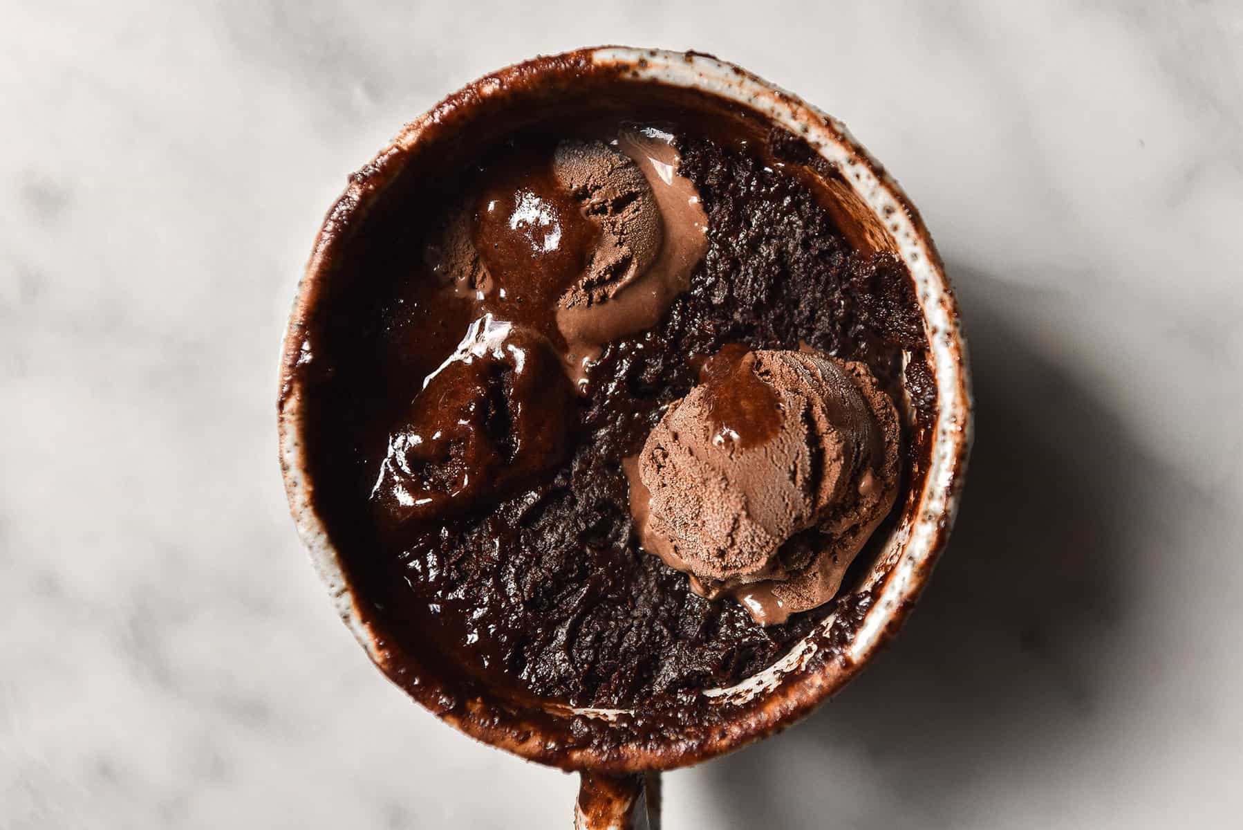 An aerial landscape view of a gluten free, vegan chocolate mug cake in a white speckled ceramic mug. The mug cake sits atop a white marble table and is topped with melting chocolate ice cream. 