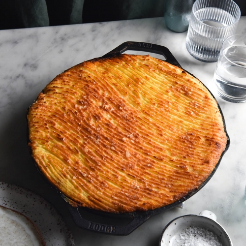 A side on view of a FODMAP friendly vegetarian Shepherd's Pie baked in a skillet. The mashed potato topping is golden brown and has a wavy pattern traced in with fork prongs. It sits atop a white marble table and is surrounded by white ceramic plates, a small white ceramic salt dish and glasses of water. The background is a dark olive piece of linen