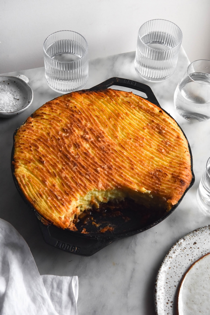 A side on view of a skillet of FODMAP friendly vegetarian Shepherd's Pie with a golden brown top with a wavy fork pattern on top. The pie sits on a white marble table and is surrounded by white ceramic plates, water glasses, a ceramic salt dish and a white linen tablecloth