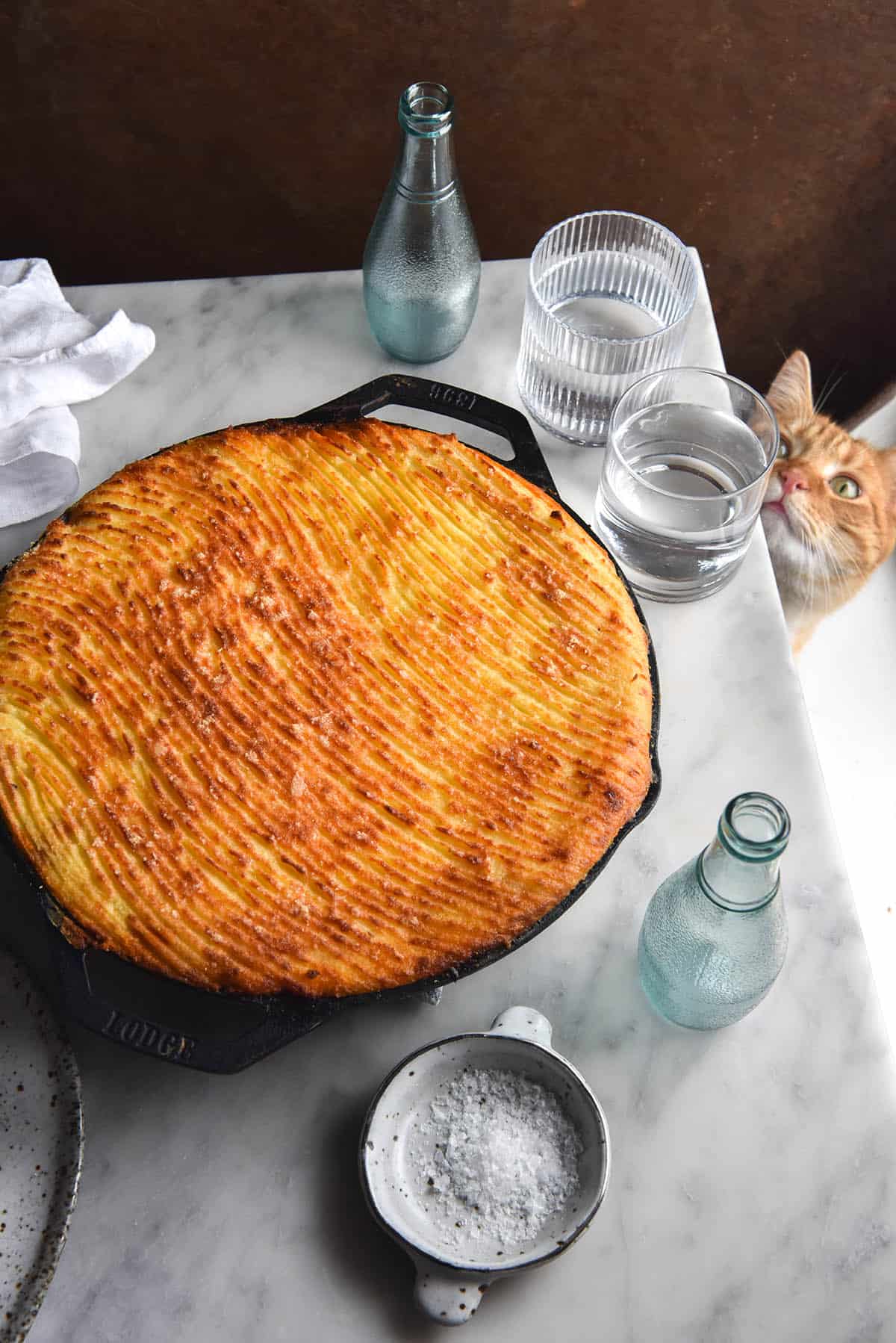 A side on view of a FODMAP friendly vegetarian Shepherd's Pie baked in a skillet. The mashed potato topping is golden brown and has a wavy pattern traced in with fork prongs. It sits atop a white marble table and is surrounded by white ceramic plates, a small white ceramic salt dish and glasses of water. The background is a dark olive piece of linen. A ginger cat pokes his head out from under the table to the right of the image.