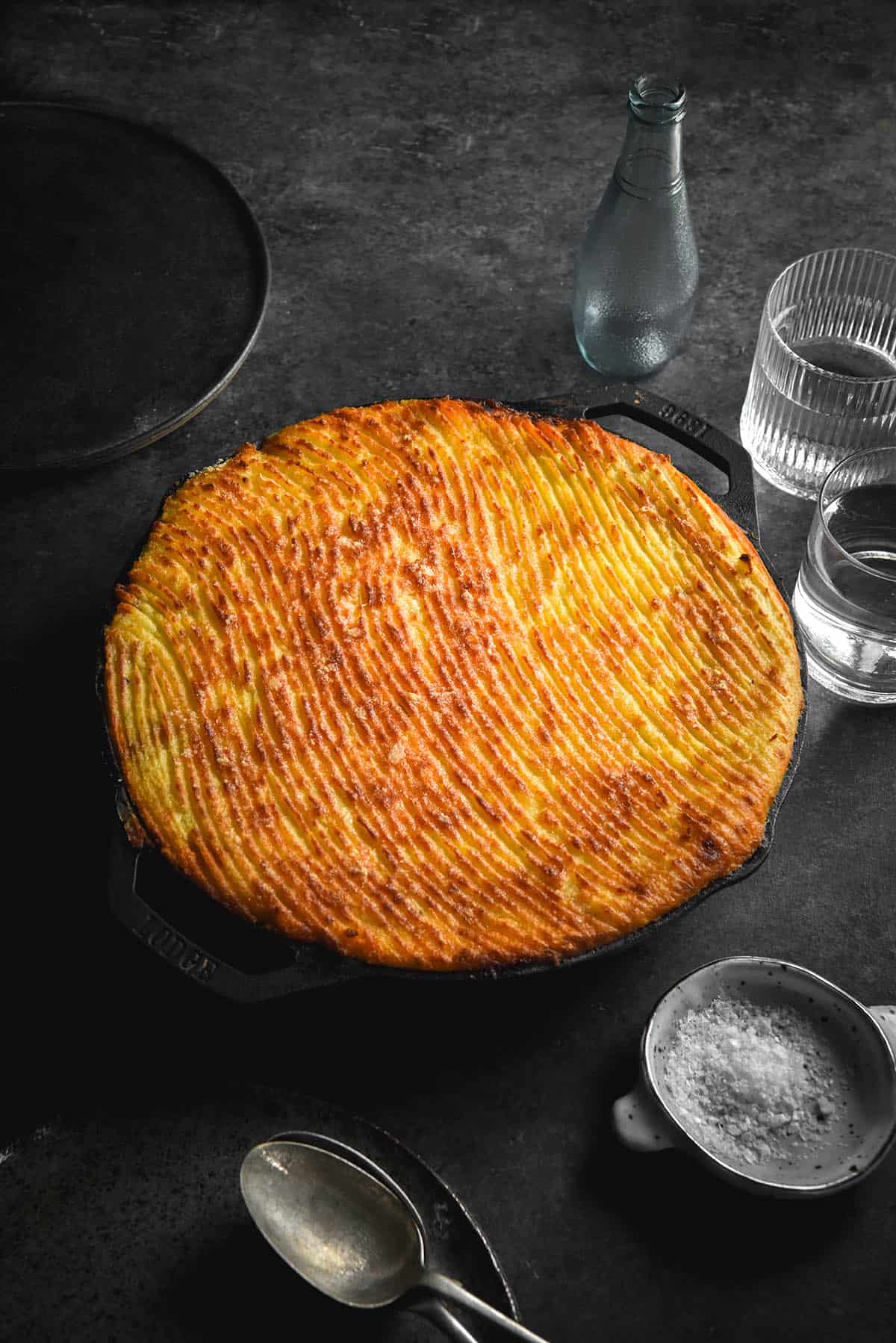 An aerial view of a FODMAP friendly vegetarian Shepherd's Pie in a skillet. The top of the Shepherd's Pie has a wavy fork pattern and is golden brown. The skillet sits atop a dark grey backdrop and is surrounded by spoons, plates, a salt dish and water glasses