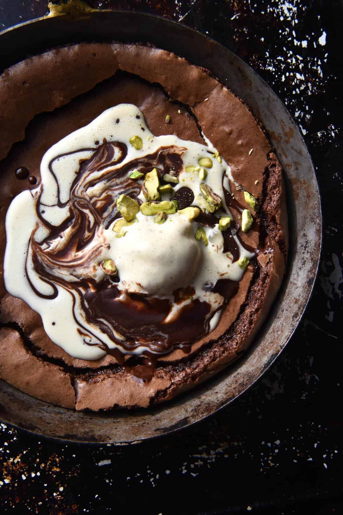 A close up aerial view of a gluten free small batch skillet brownie. The brownie is topped with melting ice cream swirled with melted chocolate and topped with some chopped pistachios. It sits atop a black mottled backdrop