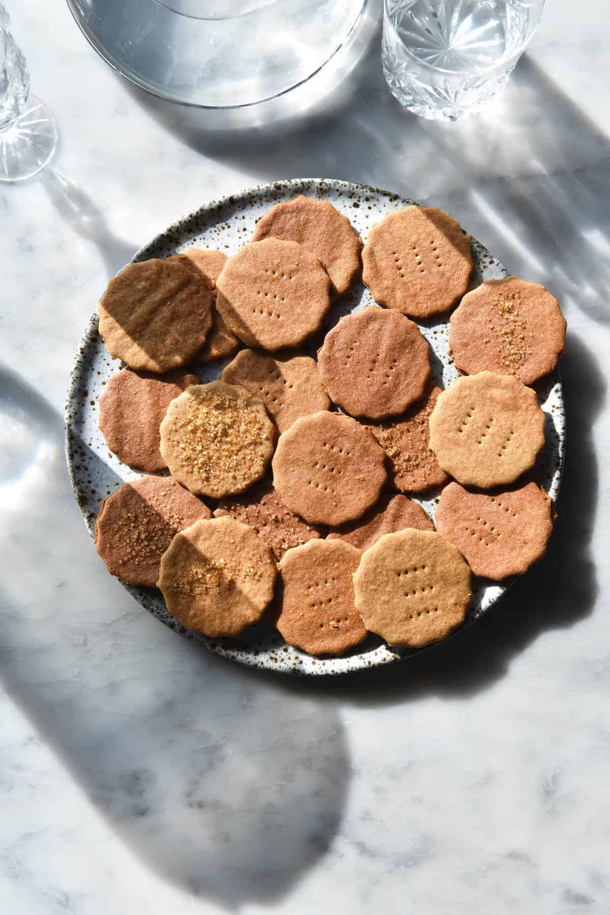 An aerial view of a white speckled ceramic plate topped with vegan gluten free Graham crackers. The crackers sit on a white marble table surrounded by glasses which project light and shadow across the image