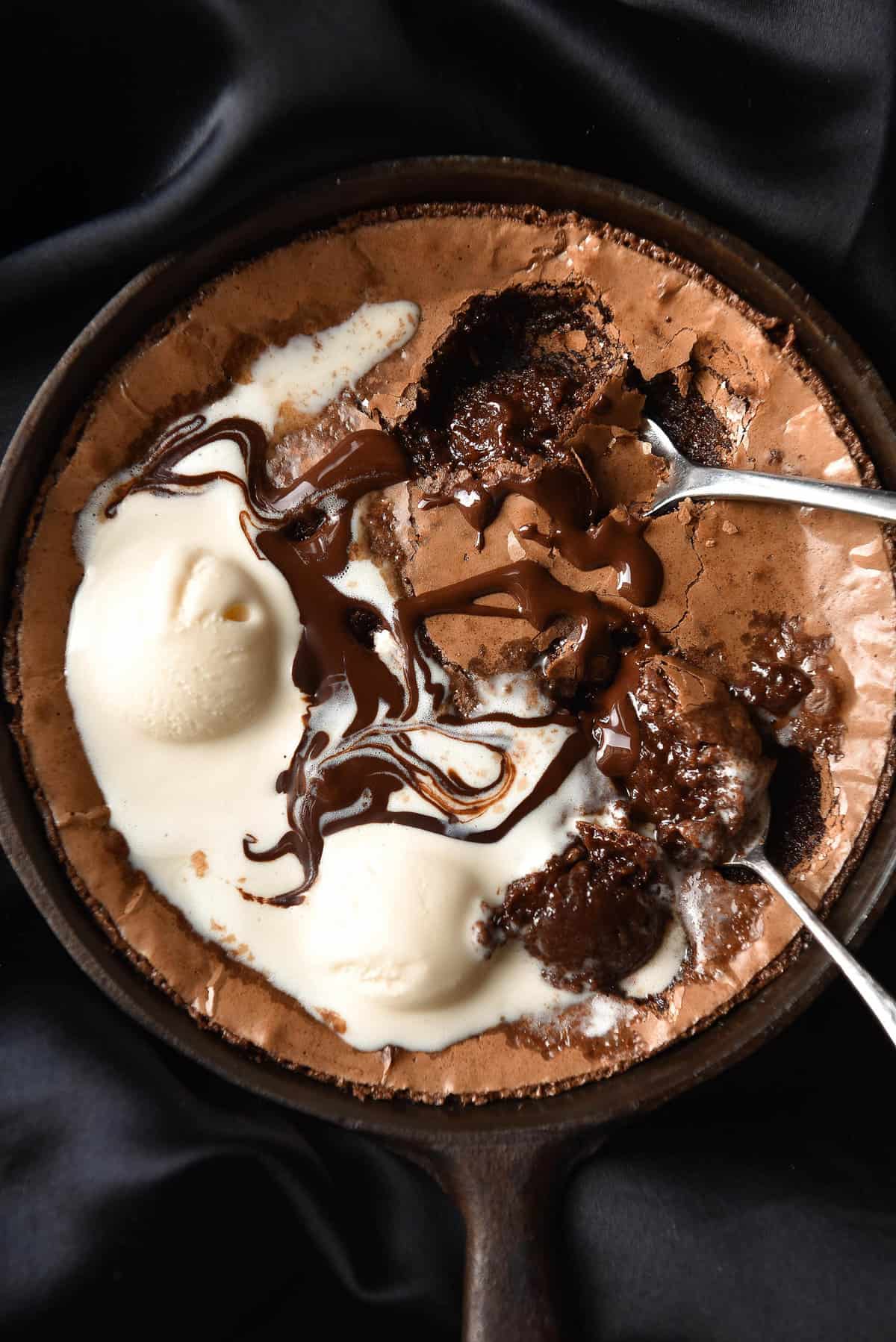 An aerial close up image of a small batch gluten free brownie in a skillet on a silky black backdrop. The brownie is topped with melting vanilla ice cream and chocolate. 