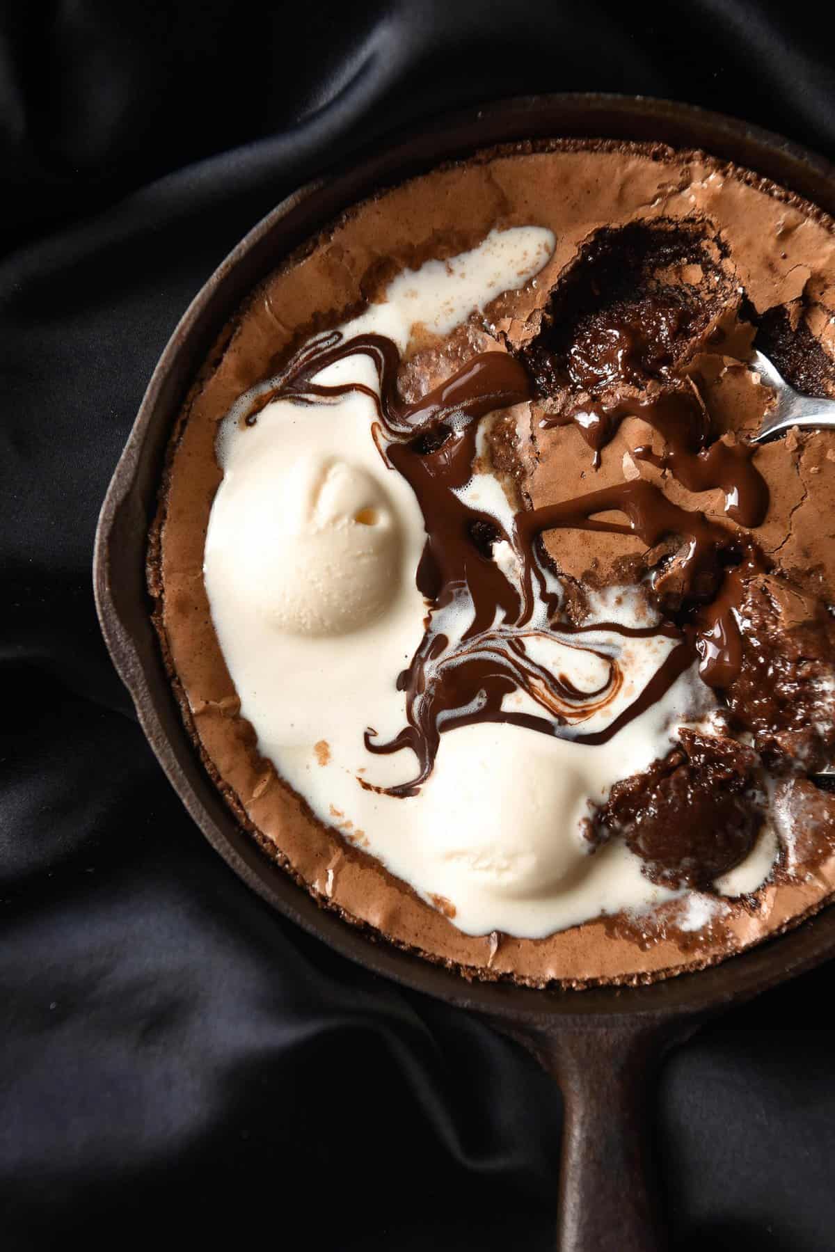 An aerial image of a skillet of gluten free brownie topped with melting vanilla ice cream and chocolate. The skillet sits atop a silky black backdrop.