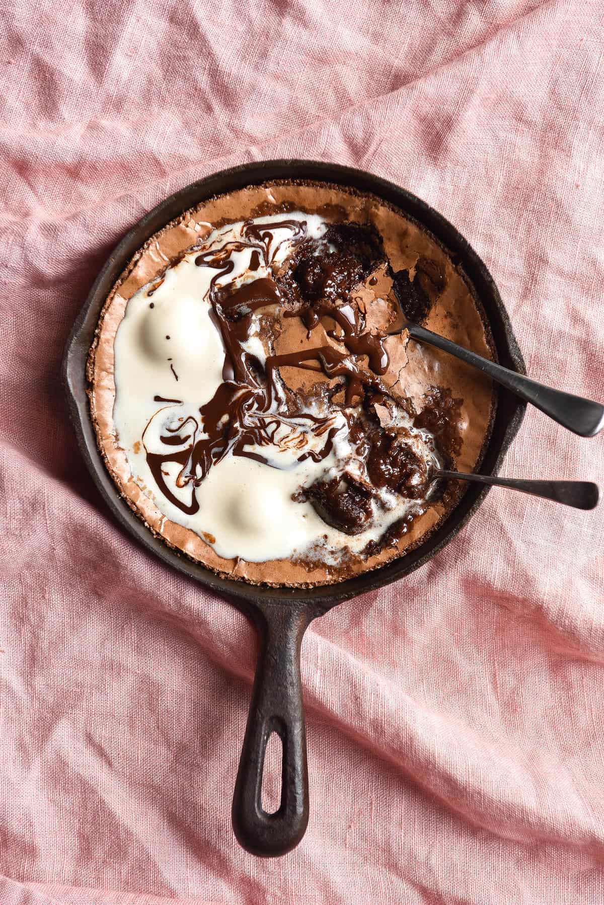 An aerial photo of a skillet of gluten free brownie on a pink linen tablecloth. The brownie is topped with melting vanilla ice cream and chocolate.
