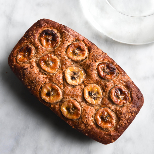 An aerial view of a loaf of gluten free banana bread atop a white marble table. The loaf is golden brown and topped with banana coins. A glass of water sits to the top right of the image, casting a light across the marble table.