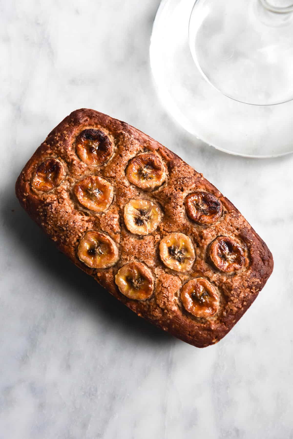 An aerial view of a loaf of gluten free banana bread atop a white marble table. The loaf is golden brown and topped with banana coins. A glass of water sits to the top right of the image, casting a light across the marble table.