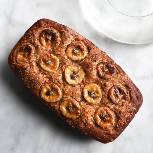 An aerial image of a loaf of gluten free dairy free banana bread on a white marble table. A sunlit bulbous vase sits to the top left of the image