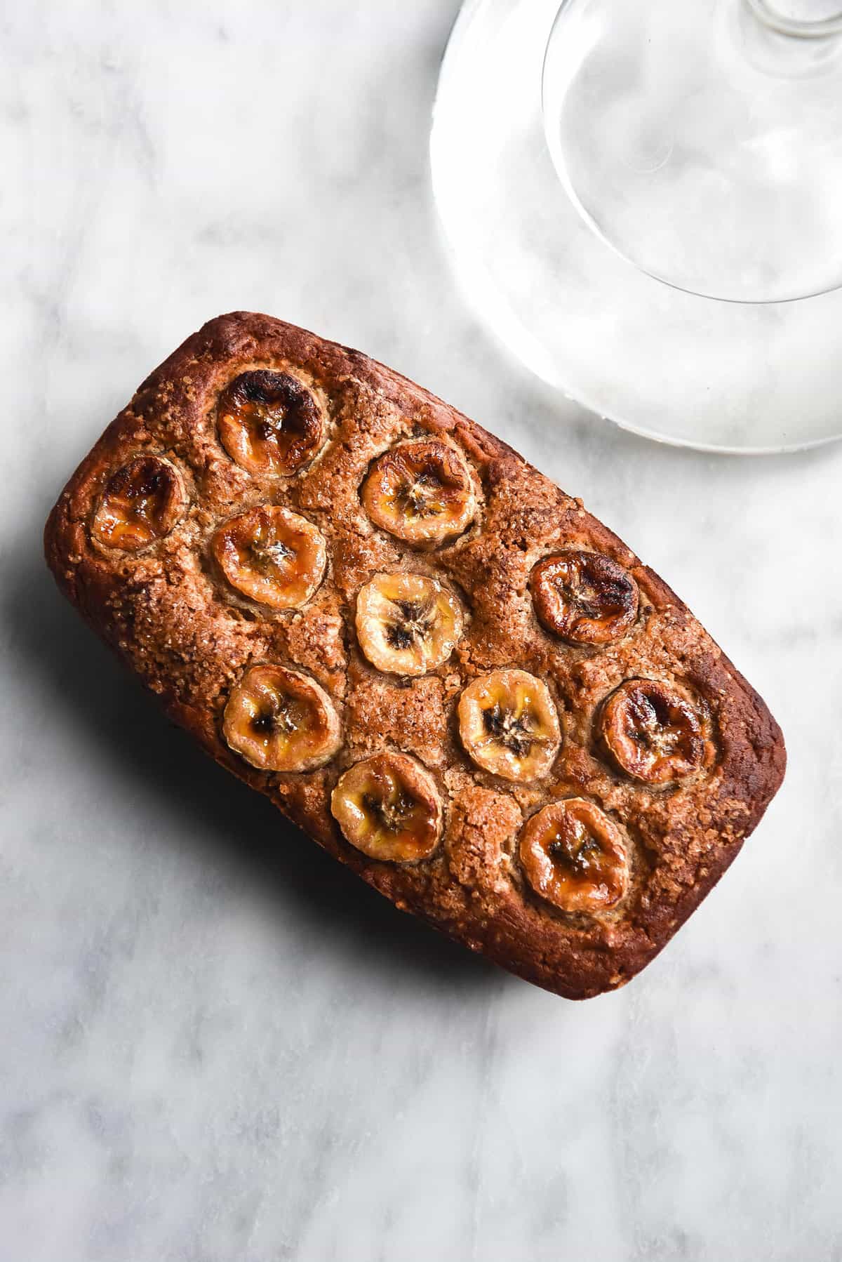 An aerial image of a loaf of gluten free dairy free banana bread on a white marble table. A sunlit bulbous vase sits to the top left of the image