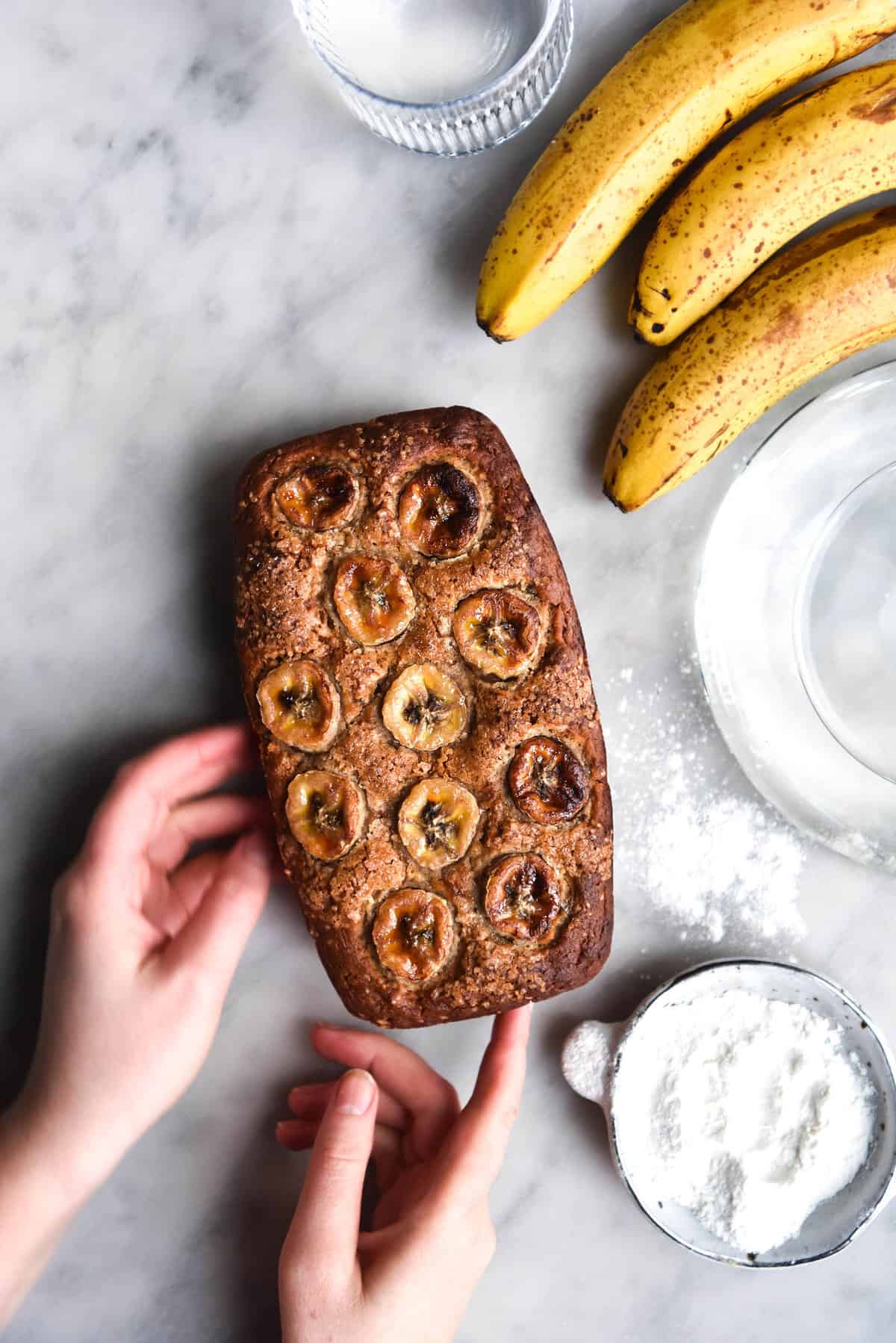 An aerial image of a loaf of gluten free dairy free banana bread on a white marble table. A sunlit bulbous vase sits to the top left of the image