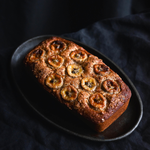 A side on moody image of a loaf of gluten free banana bread topped with banana coins. The bread sits on a steel sizzling platter on a black linen backdrop