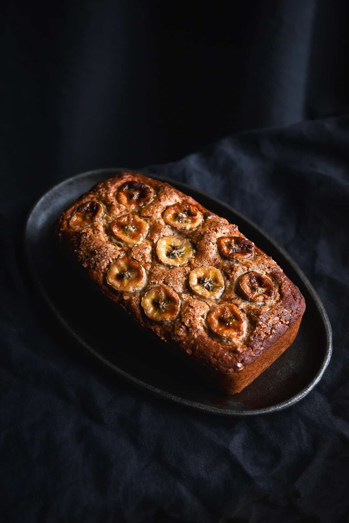 A side on moody image of a loaf of gluten free banana bread topped with banana coins. The bread sits on a steel sizzling platter on a black linen backdrop