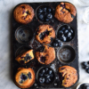 An aerial view of a muffin tray filled with gluten free blueberry muffins and extra blueberries. The tray sits atop a white marble table and is surrounded by water glasses and more blueberries