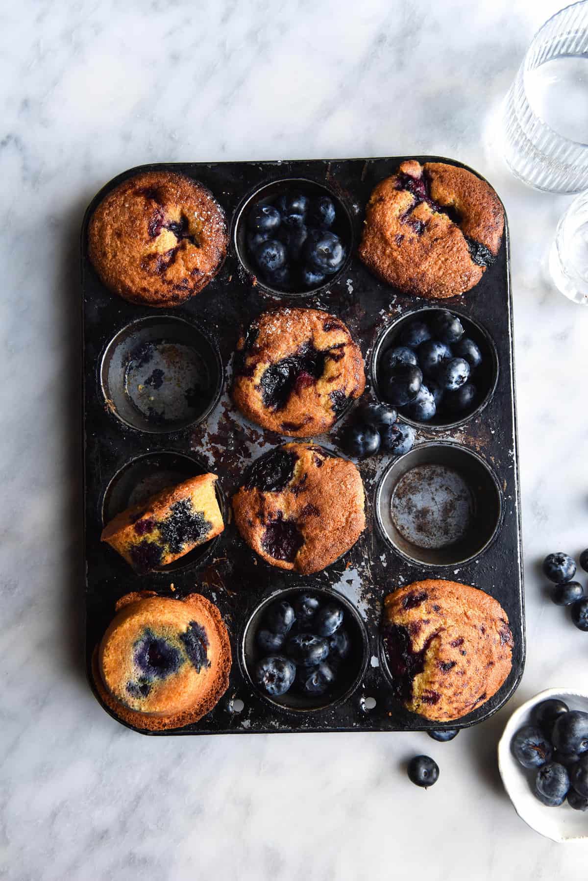 An aerial view of a muffin tray filled with gluten free blueberry muffins and extra blueberries. The tray sits atop a white marble table and is surrounded by water glasses and more blueberries