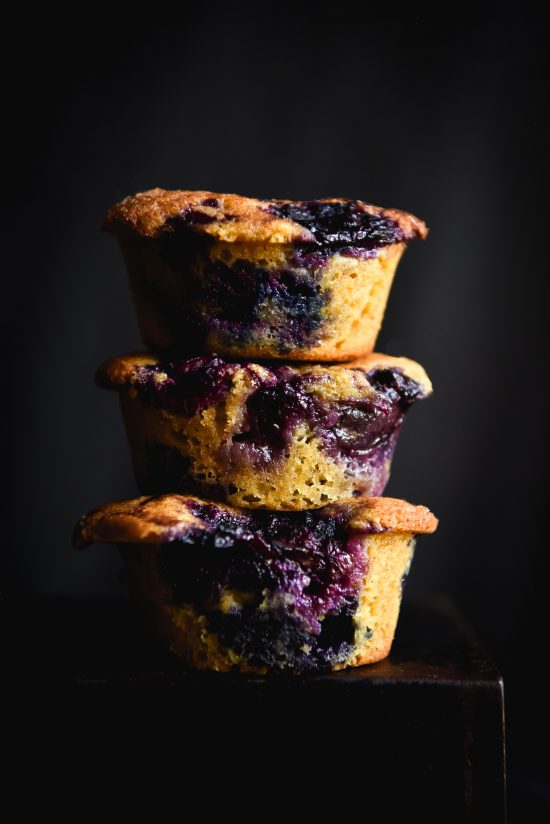 A moody side on photo of a stack of three gluten free blueberry muffins. The muffins are bursting with blueberries which contrast with the lightness of the crumb and the dark backdrop.