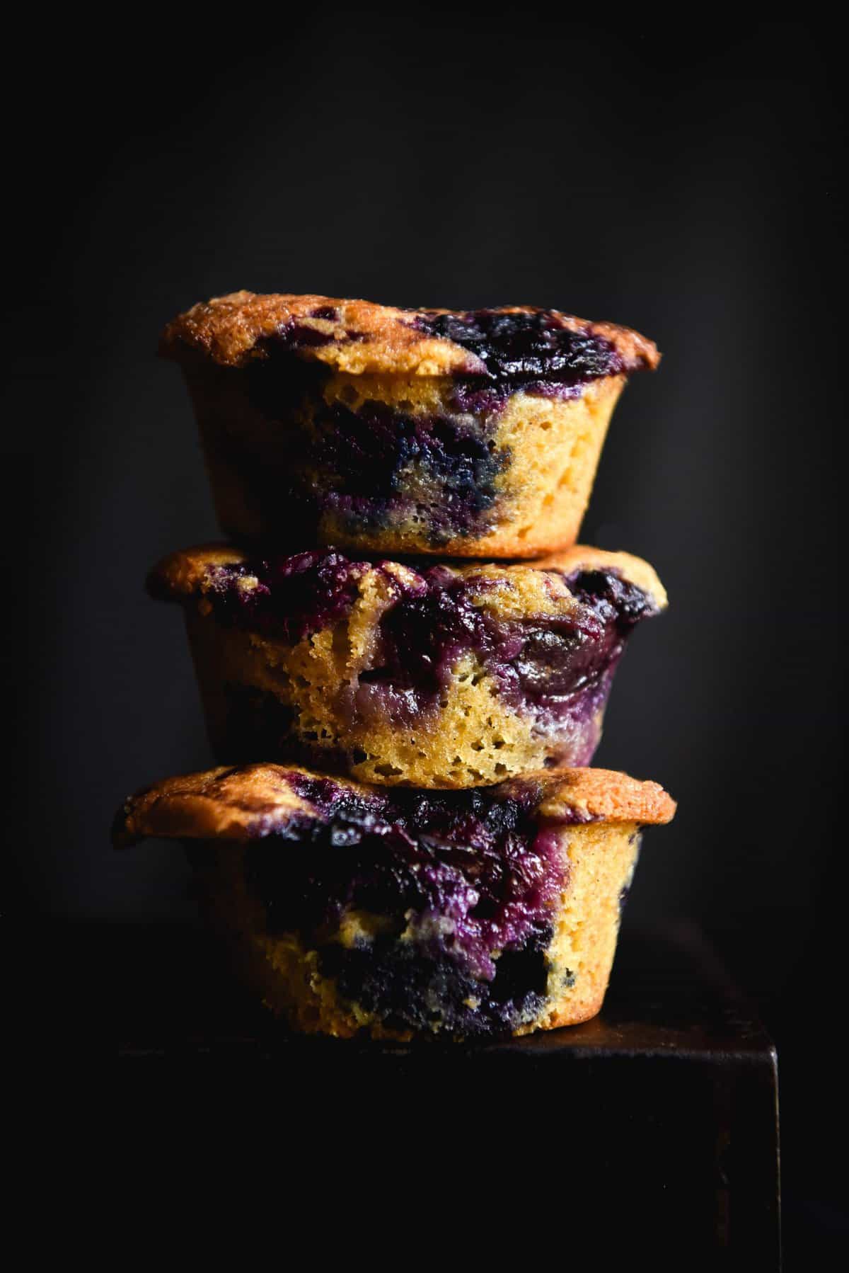 A moody side on photo of a stack of three gluten free blueberry muffins. The muffins are bursting with blueberries which contrast with the lightness of the crumb and the dark backdrop. 