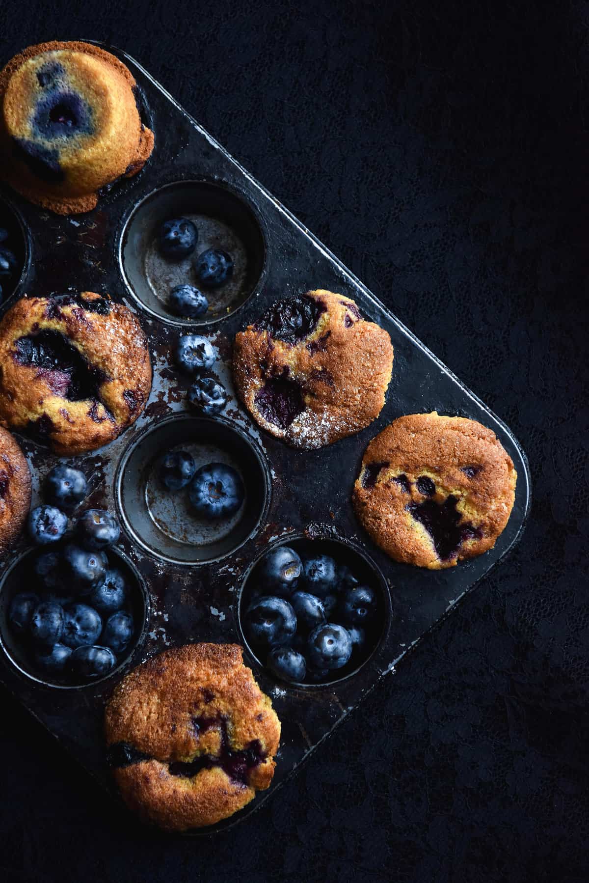 A moody aerial image of gluten free blueberry muffins in a dark muffin tray. The empty muffin holes are filled with fresh blueberries.