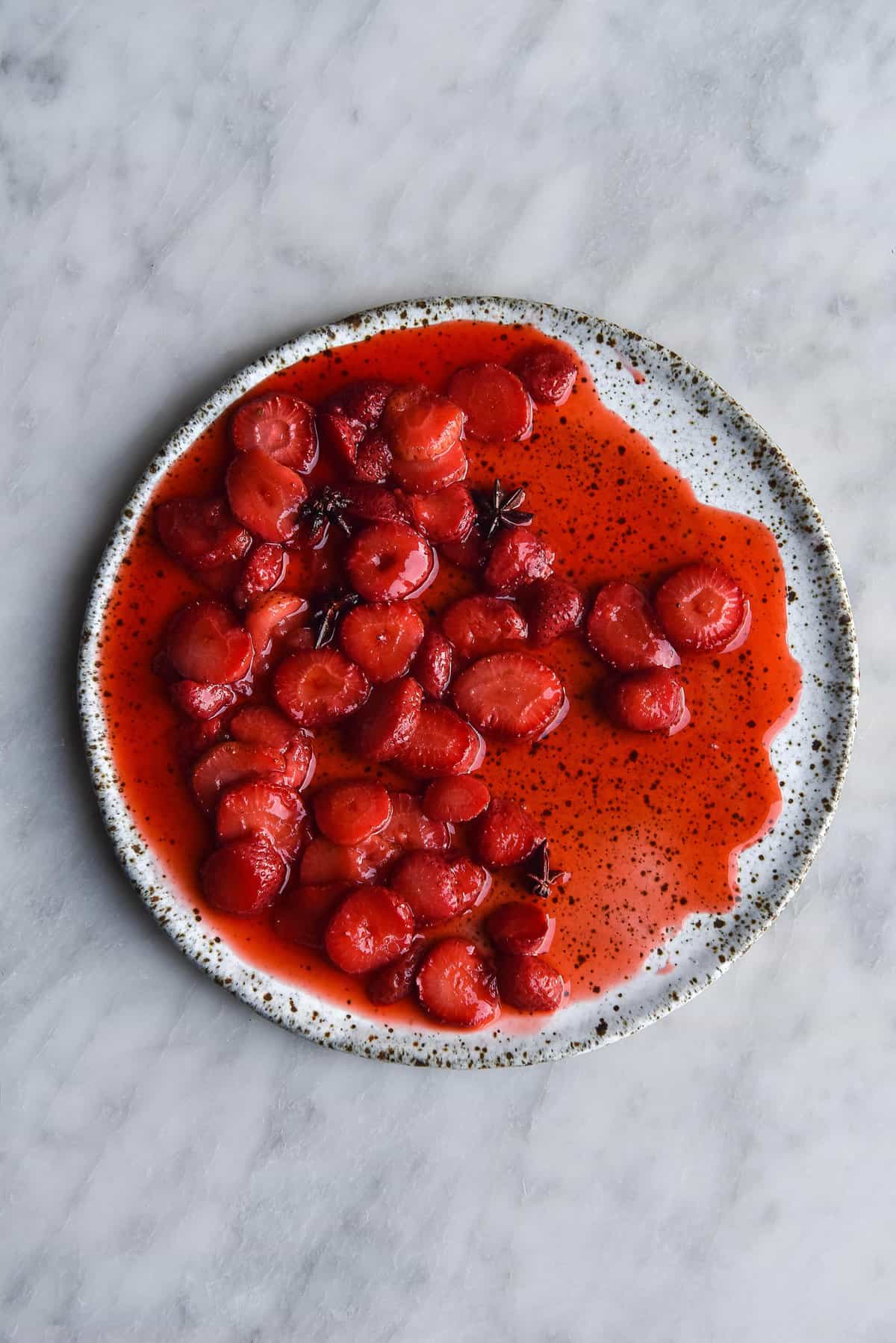 An aerial view of jammy star anise roasted strawberries on a white speckled ceramic plate atop a white marble table. 