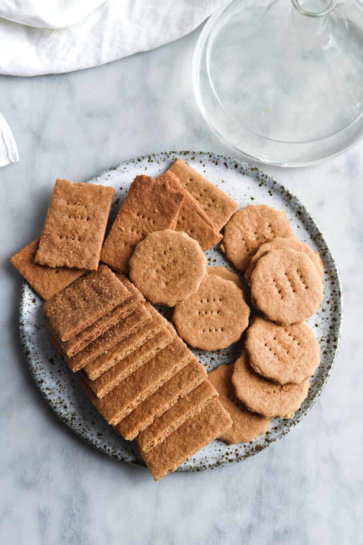 An aerial image of gluten free Graham crackers casually arranged on a white speckled ceramic plate atop a white marble table. 