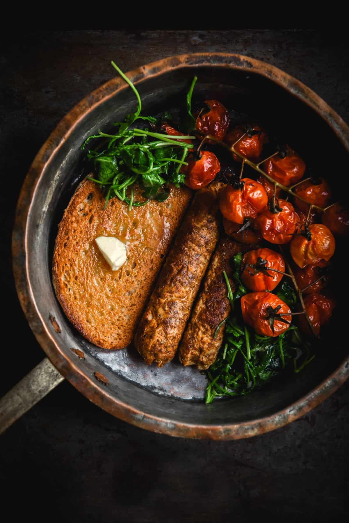 A moody aerial image of gluten free vegan sausages, roast tomatoes, wilted rocket and gluten free sourdough toast in a vintage saucepan