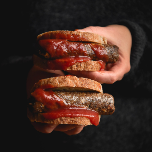 A moody side on view of a person holding out two sausage sandwiches with vegan sausages smothered in tomato sauce.