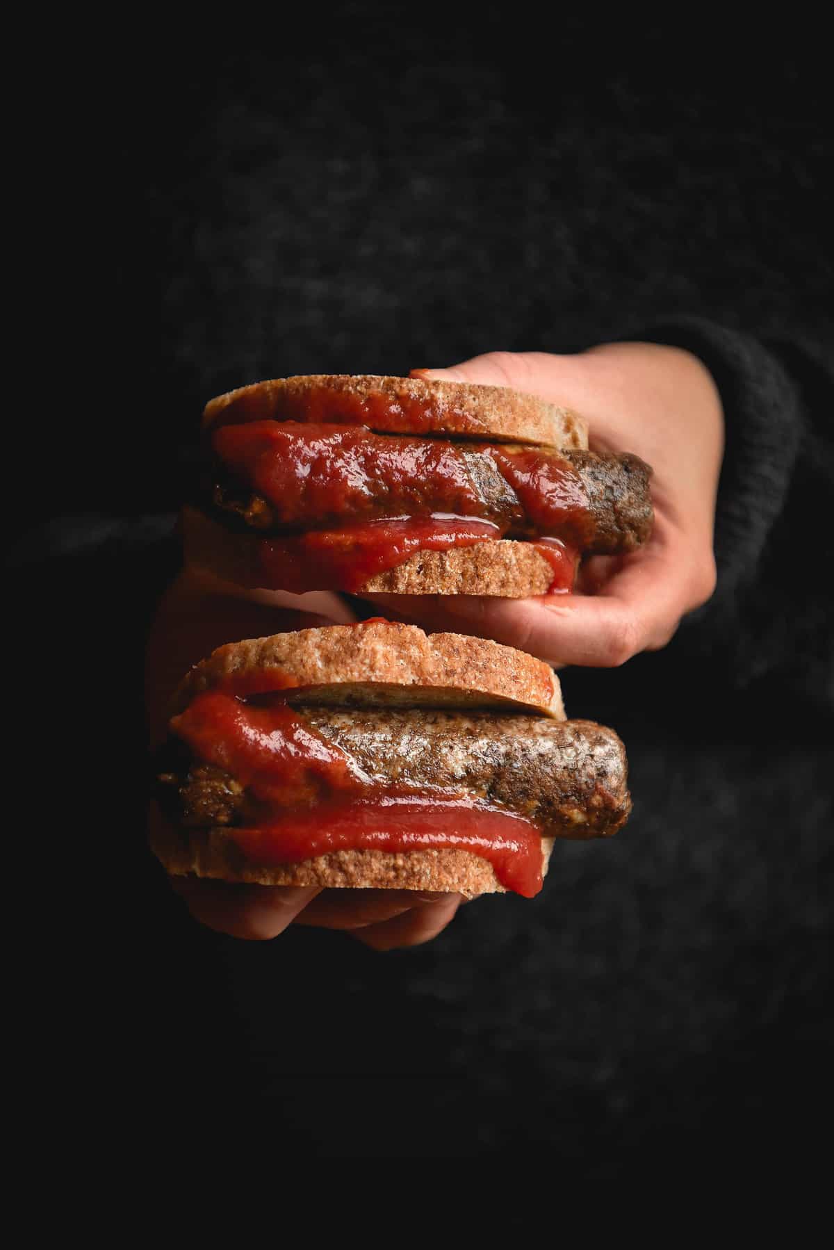 A moody side on view of a person holding out two sausage sandwiches with vegan sausages smothered in tomato sauce.