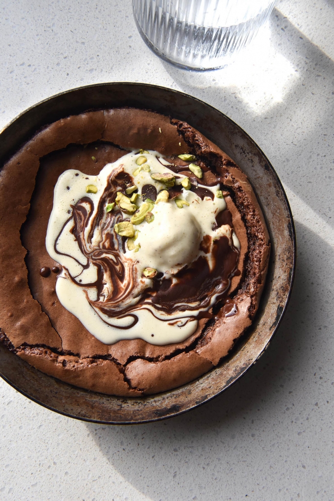 A close up aerial view of a gluten free small batch skillet brownie. The brownie is topped with melting ice cream swirled with melted chocolate and some chopped pistachios. It sits atop a white kitchen counter and a light flooded glass of water sits to the top right of the image.