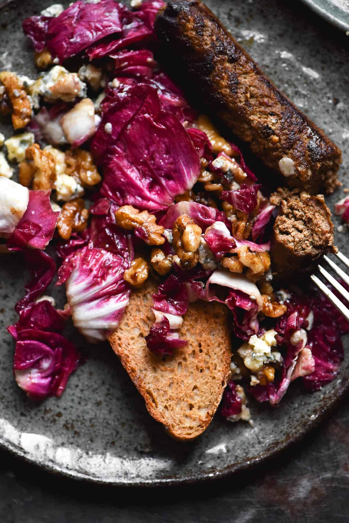 An aerial close up view of a radicchio, walnut and blue cheese salad served with gluten free sourdough and vegan sausages. The meal is casually arranged on a dark blue ceramic plate and sits on a steel blue backdrop