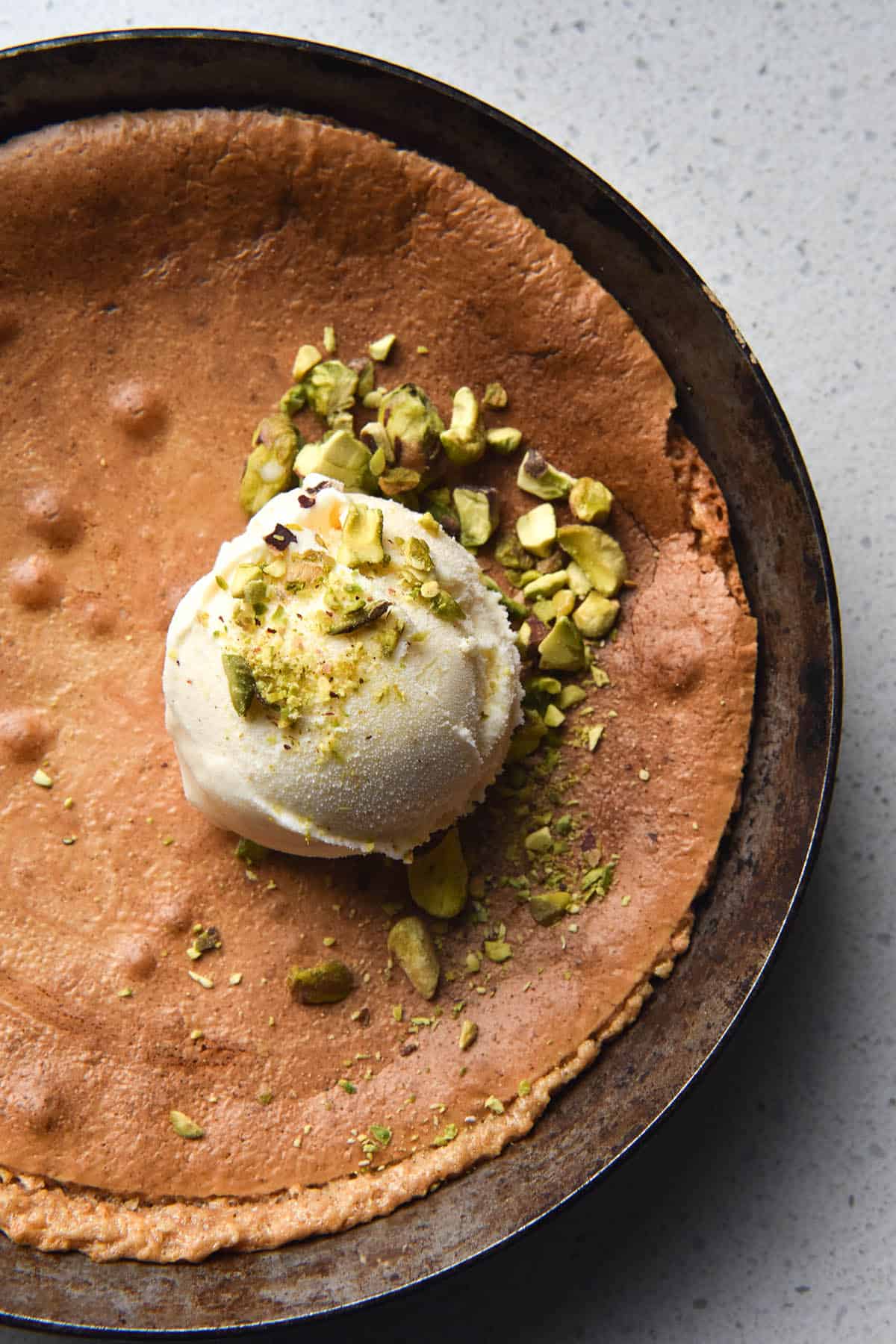 An aerial, close up image of a white chocolate small batch skillet brownie. The brownie is topped with a scoop of vanilla ice cream and chopped pistachios. It sits atop a white table top.