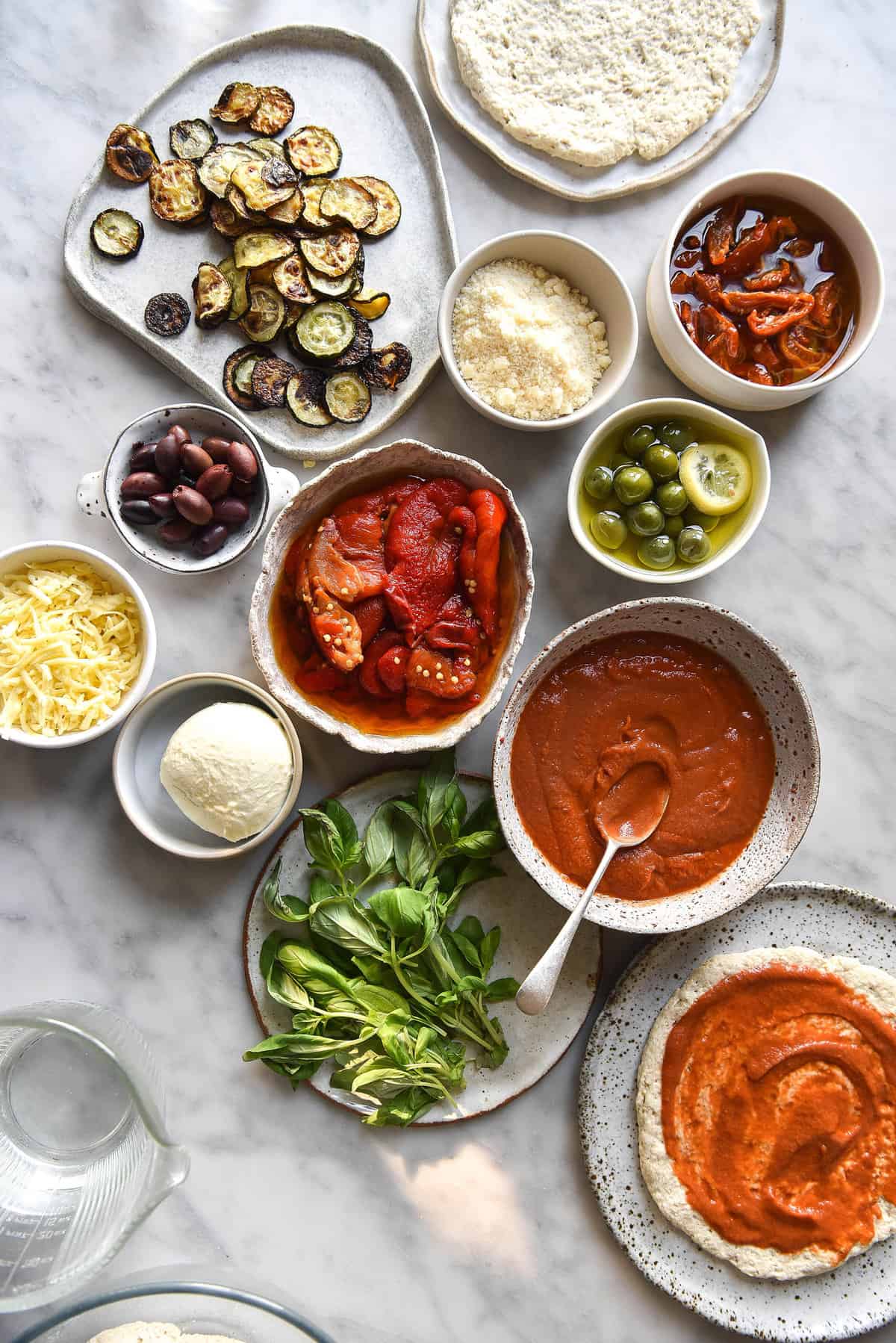 An array of vegetarian pizza ingredients in variously sized white ceramic bowls on a white marble backdrop