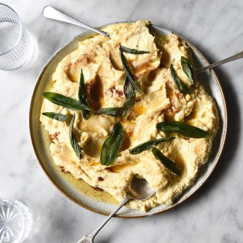 Brown butter mashed potatoes on a white ceramic plate against a white marble table. The mashed potato is topped with crispy sage leaves and surrounded by sunlit glasses of water