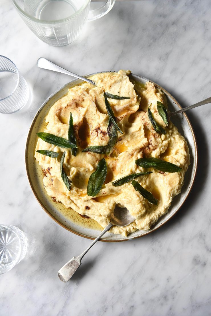 Brown butter mashed potatoes on a white ceramic plate against a white marble table. The mashed potato is topped with crispy sage leaves and surrounded by sunlit glasses of water