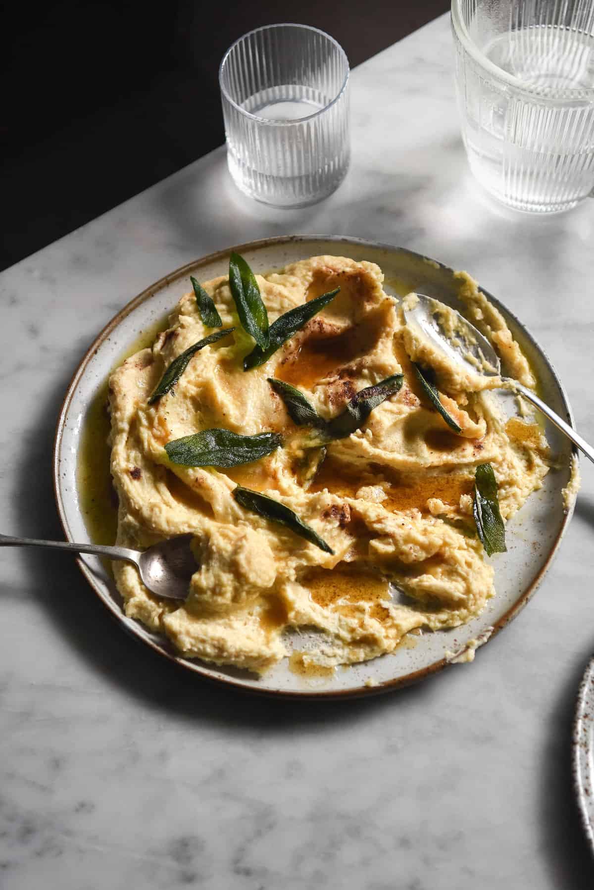 A moody image of brown butter mashed potatoes topped with crispy sage on a white marble table. 