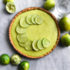An aerial image of a gluten free lime curd tart atop a white marble table. The tart is topped with thin slices of lime and surrounded by extra squeezed and whole limes