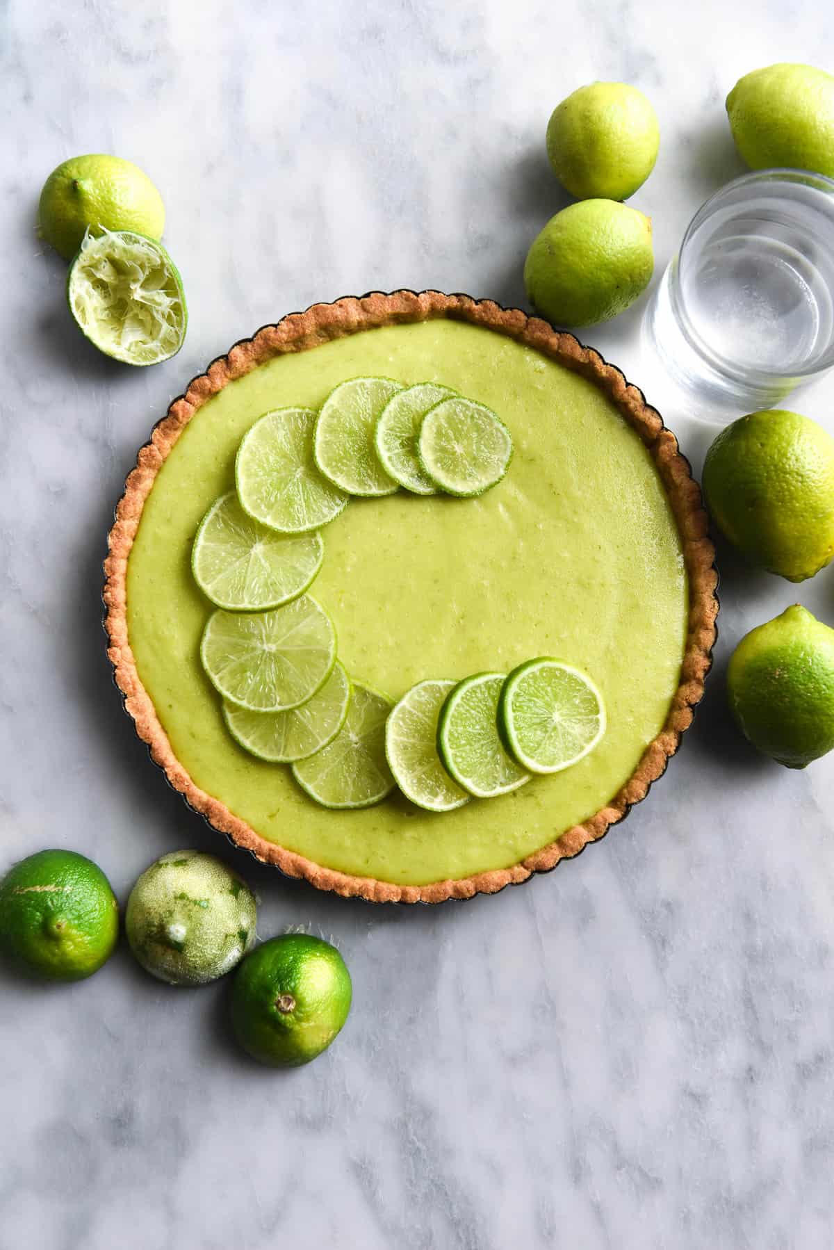 An aerial image of a gluten free lime curd tart atop a white marble table. The tart is topped with thin slices of lime and surrounded by extra squeezed and whole limes