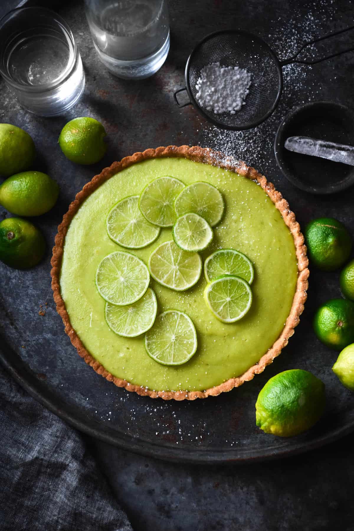 An aerial view of a gluten free lime curd top on a medium blue steel backdrop. The tart is topped with fresh slices of lime and surrounded by extra squeezed and fresh limes