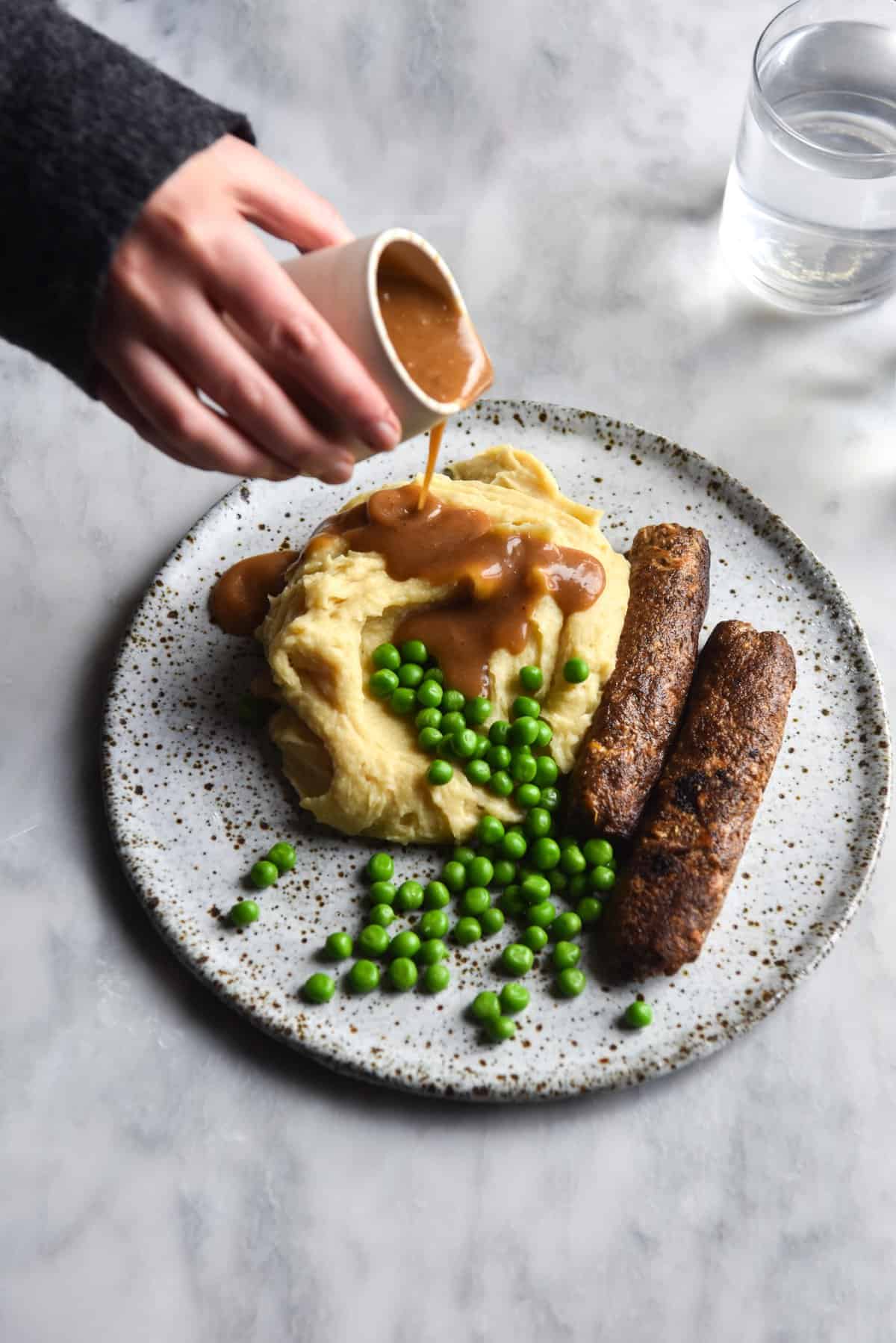 An aerial image of a white ceramic plate topped with mashed potatoes, vegan sausages and peas. The plate sits on a white marble table with a water glass in the back right corner. A hand extends out to pour gravy over the mashed potatoes from the top left corner of the image. 