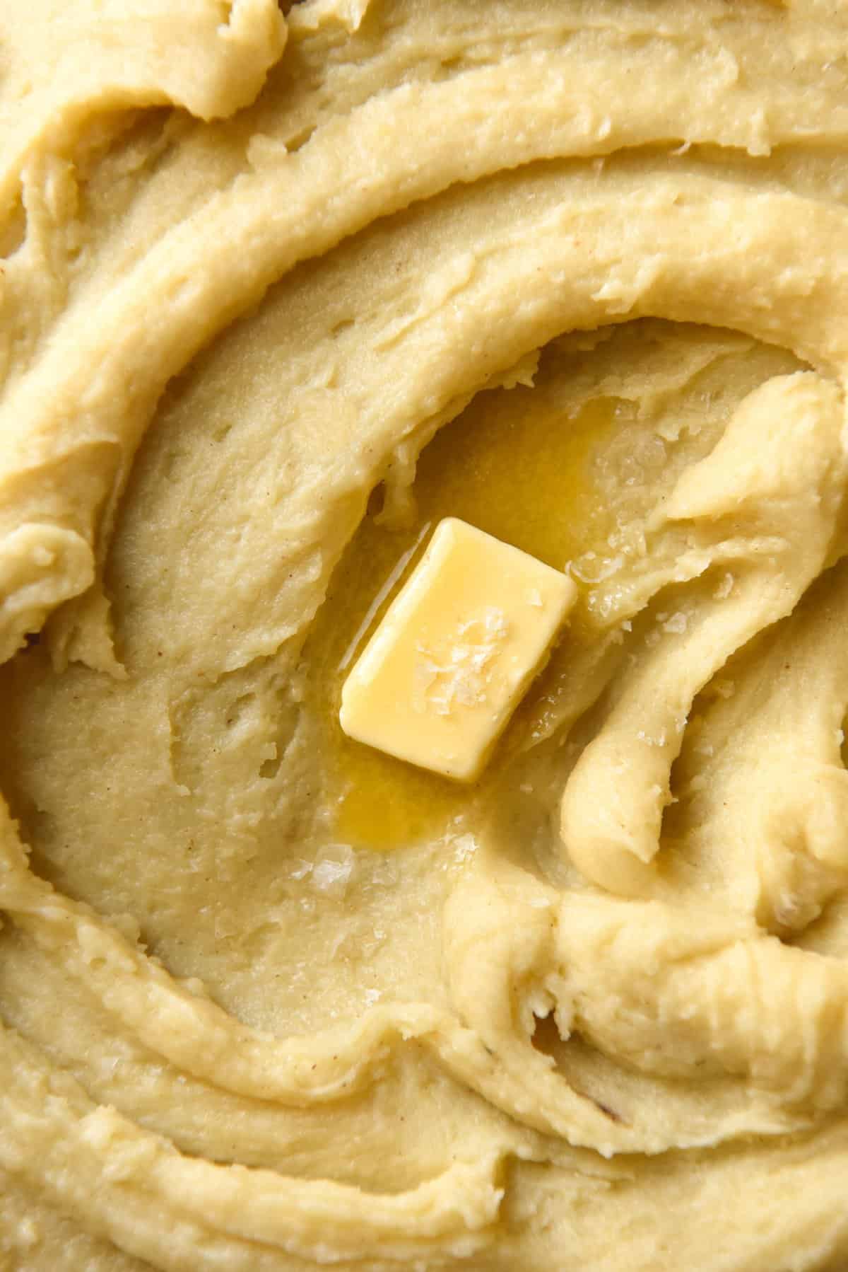 A close up aerial view of a plate of browned butter mashed potato. The mashed potato has swoops and swirls and a piece of melting butter topped with sea salt flakes sits in the centre