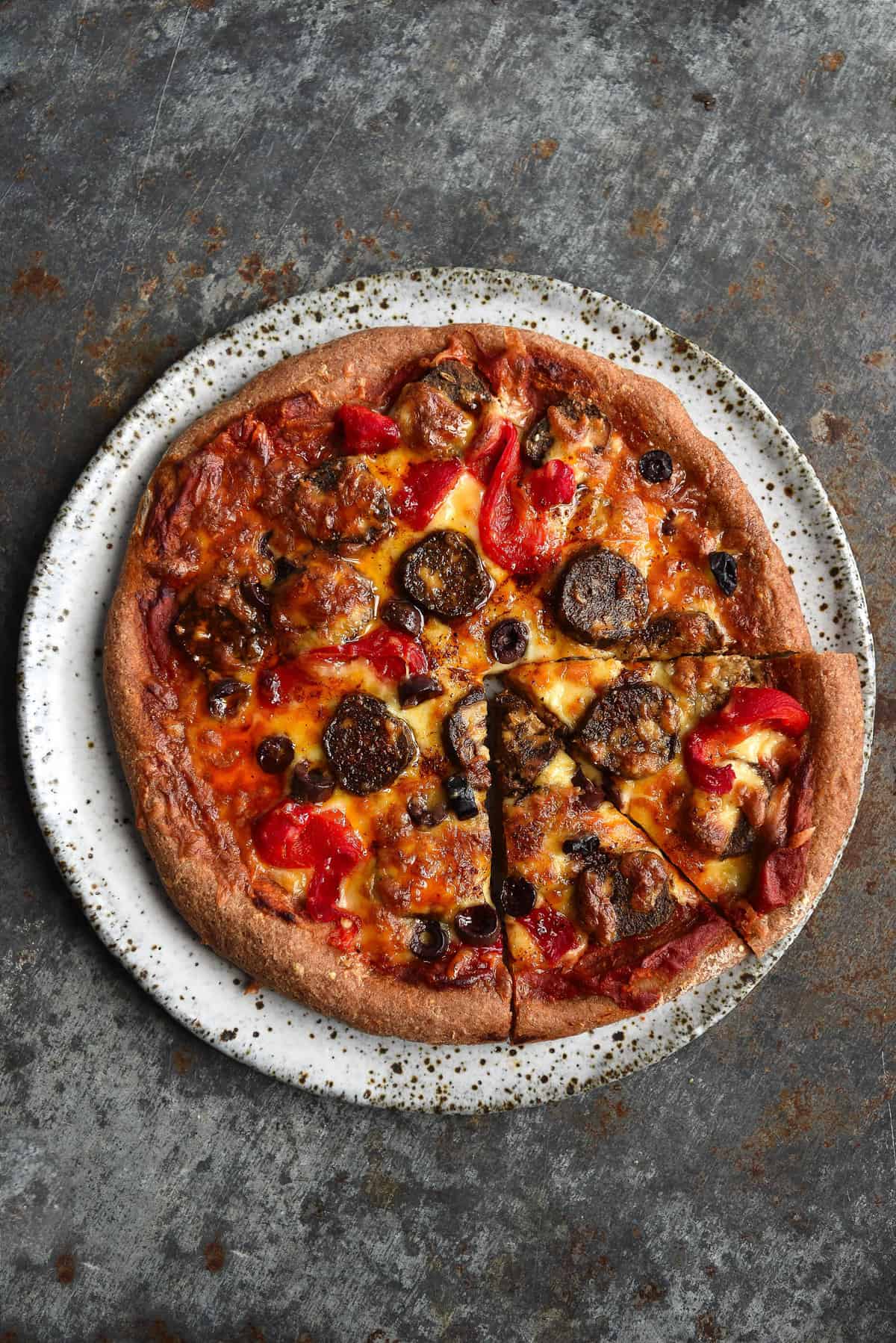An aerial image of a gluten free pizza topped with low FODMAP pizza sauce, vegan sausage and red capsicums. The pizza sits on a white speckled ceramic plate atop a steel backdrop.