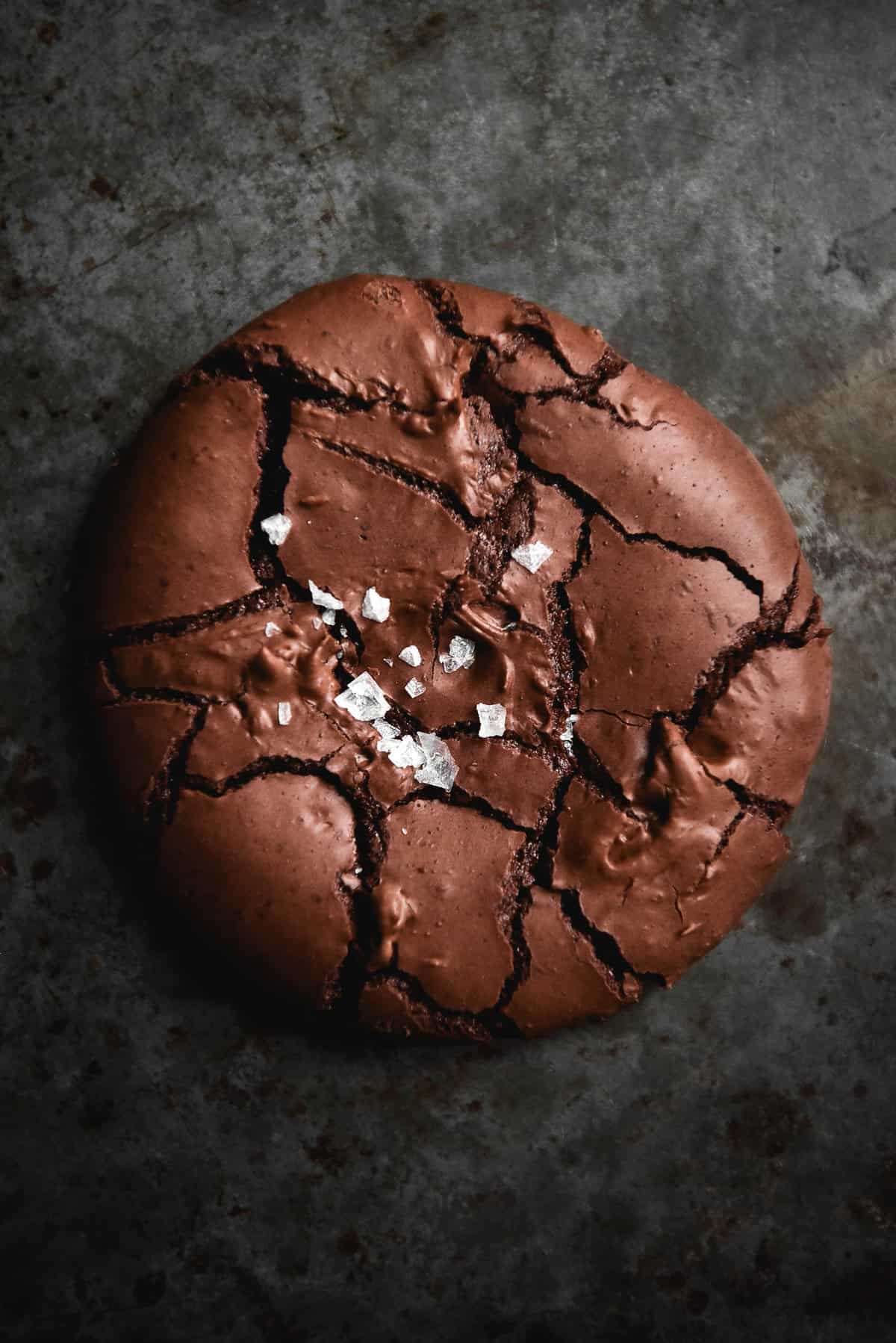 An aerial close up image of a flourless brownie cookie atop a dark grey backdrop. The brownie cookie is shiny and crackled and topped with sea salt flakes that contrast with the deep chocolate colour of the cookie