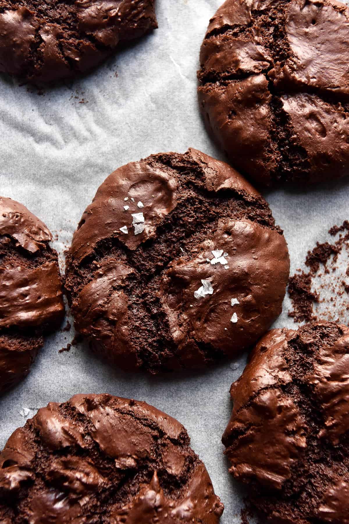 An aerial close up image of gluten free vegan brownie cookies casually arranged on a lined baking sheet. The cookies are fudgy and topped with a small sprinkle of sea salt flakes. 