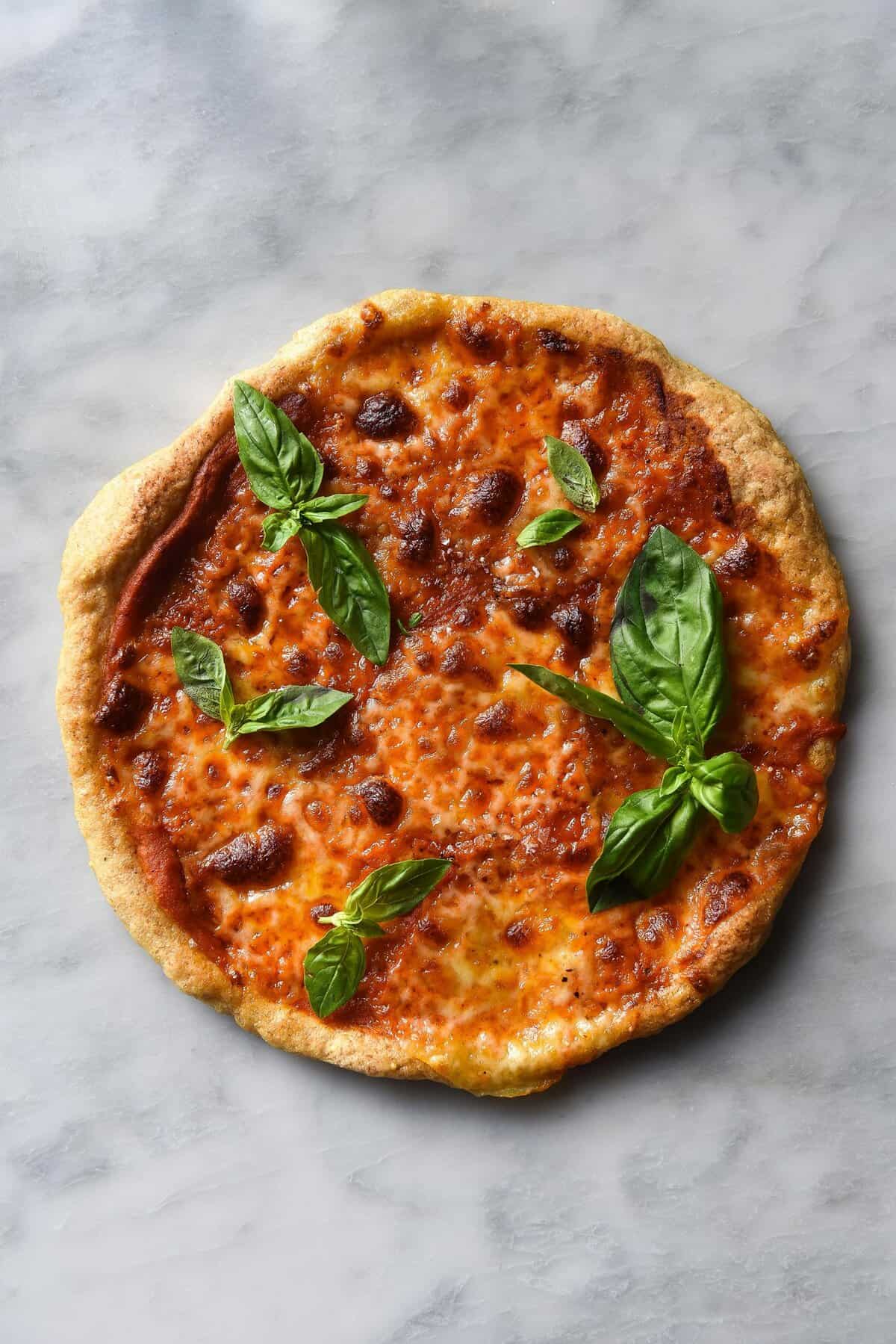 An aerial view of a gluten free pizza topped with low friendly pizza sauce, melty cheese and basil leaves. The pizza sits atop a white marble table.  