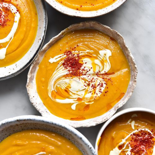 An aerial image of white ceramic bowls filled with FODMAP friendly pumpkin soup on a white marble table. The bowls of soup are topped with cream and red chilli flakes.