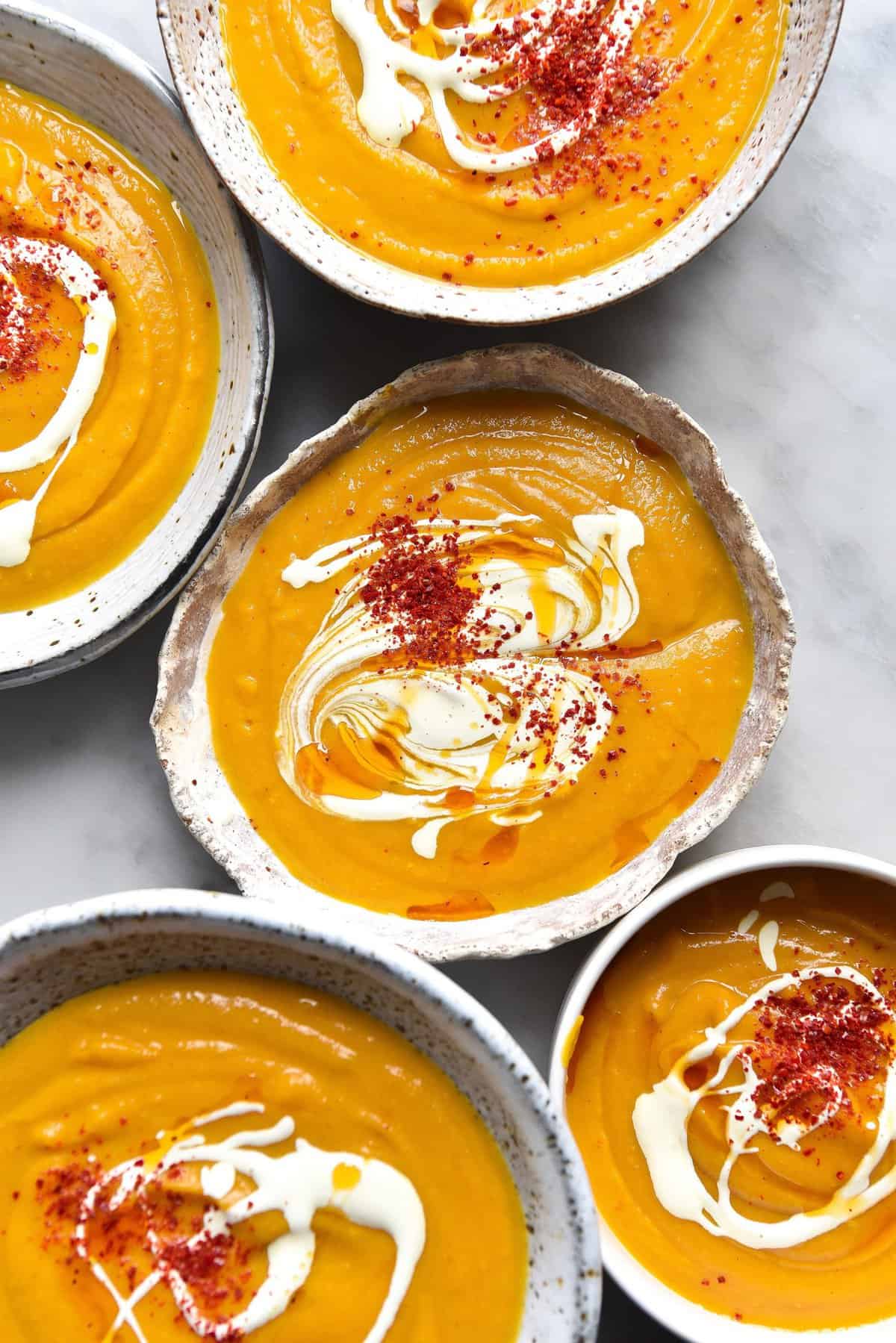 An aerial image of white ceramic bowls filled with FODMAP friendly pumpkin soup on a white marble table. The bowls of soup are topped with cream and red chilli flakes. 