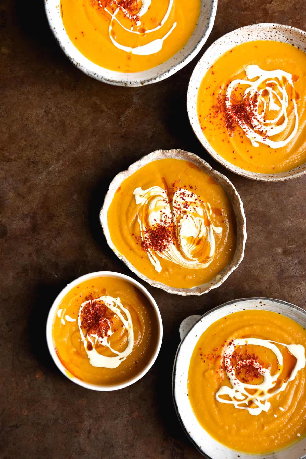 An aerial image of 5 ceramic white bowls filled with pumpkin soup atop a rusty backdrop. The soup bowls are topped with swirls of cream and chilli flakes. 