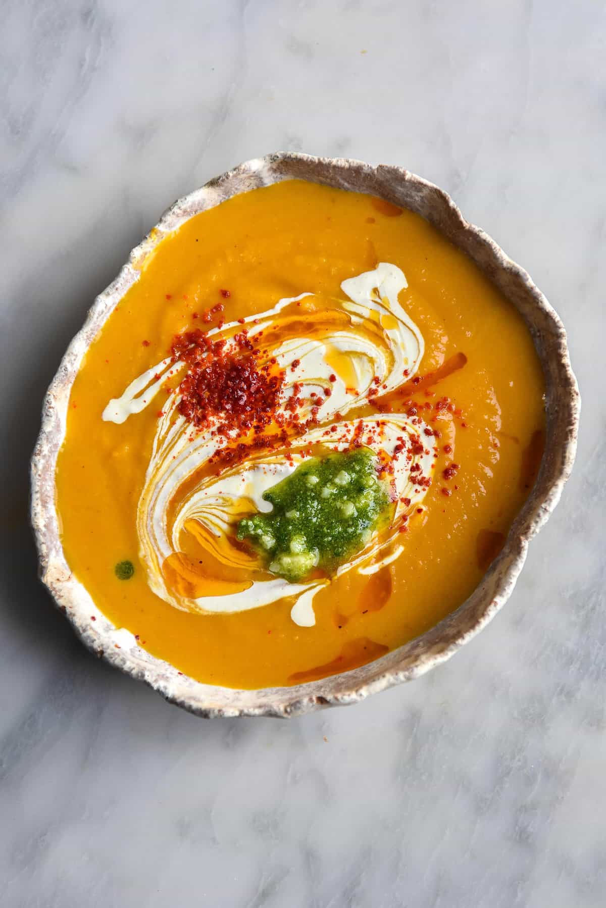 An aerial image of a white ceramic bowl of pumpkin soup topped with swirls of cream, chilli flakes and pesto. The bowl sits atop a white marble table. 