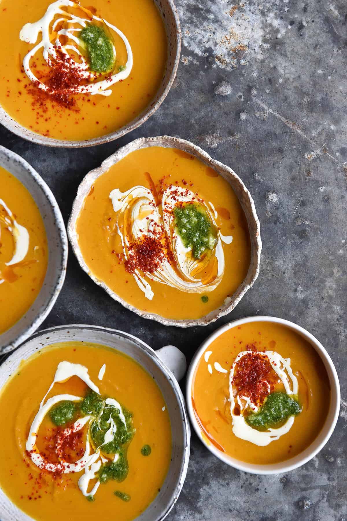 An aerial view of five bowls of FODMAP friendly pumpkin soup topped with a swirl of cream, pesto and chilli flakes. The bowls sit casually arranged atop a mottled grey blue backdrop