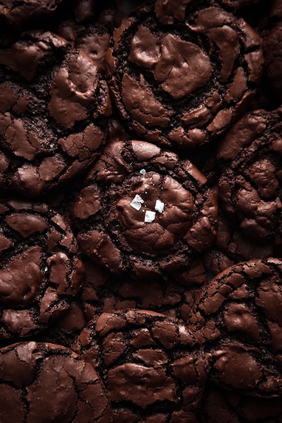 An aerial, moody photo of a stack of vegan brownie cookies that are egg free, gluten free and made without xanthan gum. The cookies have shiny, crackled tops and the cookie on the top of the stack at the centre is sprinkled with sea salt flakes