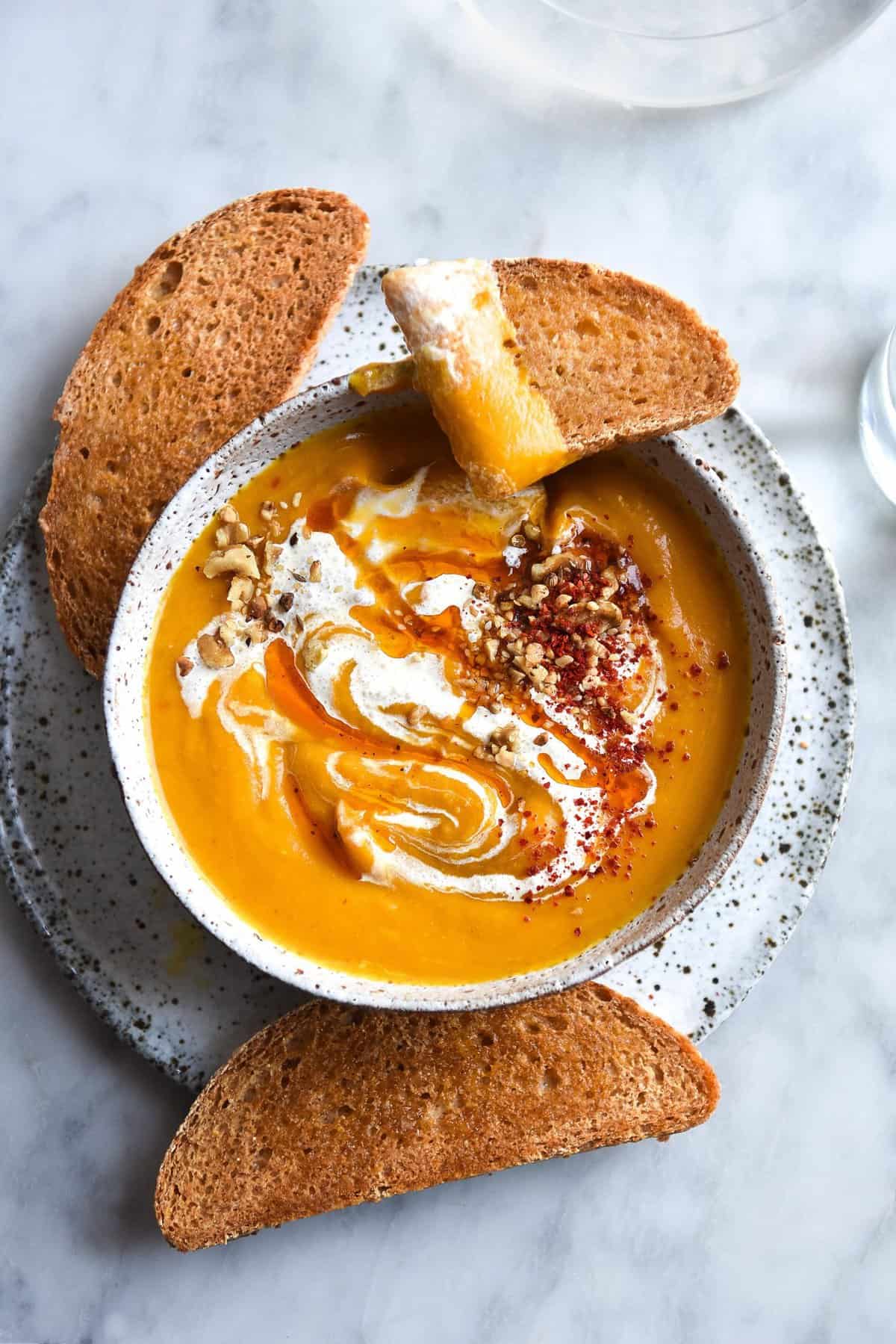 A white speckled ceramic bowl of pumpkin soup on a white speckled plate atop a white marble table. Slices of sourdough sits to the bottom and top of the soup, which has been topped with cream, chilli oil and dukkah.